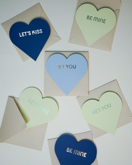 Letterpress Valentine blue and green greeting cards in the shape of hearts with foiled lettering saying Let's Kiss, Be Mine, and Hey You, Link