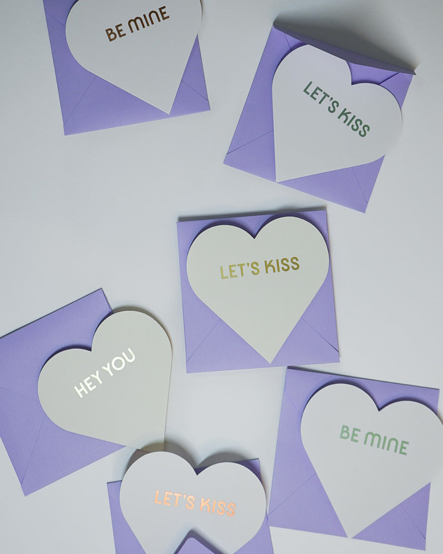 Letterpress Valentine white greeting cards in the shape of hearts with foiled lettering saying Let's Kiss, Be Mine, and Hey You with purple envelopes, Link